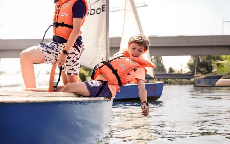 Zwei Kinder mit orangefarbenen Schwimmwesten sitzen auf einem kleinen Segelboot; eines lehnt sich über den Rand und berührt mit seiner Hand das Wasser.