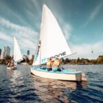 Zwei kleine Segelboote mit Personen in Schwimmwesten auf einem See, im Hintergrund sind die Skyline der Stadt und Bäume unter blauem Himmel zu sehen.