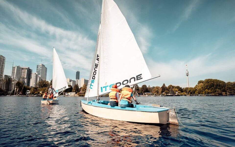 Zwei kleine Segelboote mit Personen in Schwimmwesten auf einem See, im Hintergrund sind die Skyline der Stadt und Bäume unter blauem Himmel zu sehen.