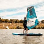 Eine Person, die eine Schwimmweste trägt, surft auf einem ruhigen Gewässer mit Bäumen und Menschen auf dem grasbewachsenen Ufer im Hintergrund.