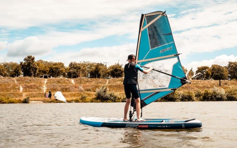 Eine Person, die eine Schwimmweste trägt, surft auf einem ruhigen Gewässer mit Bäumen und Menschen auf dem grasbewachsenen Ufer im Hintergrund.