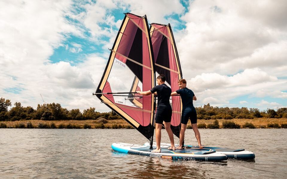 Zwei Personen stehen auf einem Paddelbrett mit großen Windsurfsegeln auf einem ruhigen Gewässer, mit Bäumen und einem bewölkten Himmel im Hintergrund.