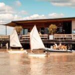 Zwei kleine Segelboote mit Kindern in Schwimmwesten und ein aufblasbares Motorboot befinden sich auf einem Fluss in der Nähe eines modernen Gebäudes, das von einer Terrasse aus beobachtet wird.