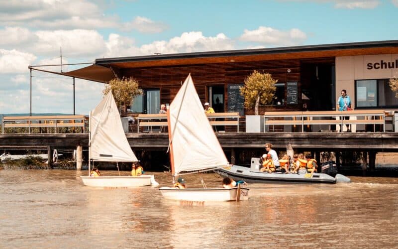 Zwei kleine Segelboote mit Kindern in Schwimmwesten und ein aufblasbares Motorboot befinden sich auf einem Fluss in der Nähe eines modernen Gebäudes, das von einer Terrasse aus beobachtet wird.