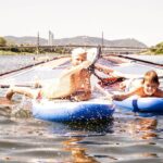 Zwei Jungen auf einem aufblasbaren Windsurfbrett und einem blauen Schwimmer genießen das Planschen im Wasser an einem sonnigen Tag, mit einer Brücke und Hügeln im Hintergrund.