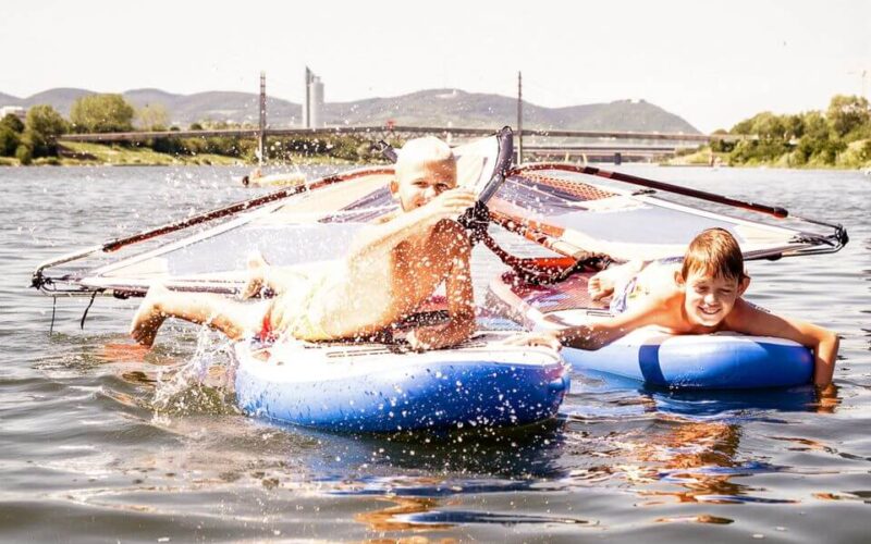 Zwei Jungen auf einem aufblasbaren Windsurfbrett und einem blauen Schwimmer genießen das Planschen im Wasser an einem sonnigen Tag, mit einer Brücke und Hügeln im Hintergrund.