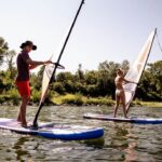 Zwei Personen surfen auf Paddelbrettern auf einem See, der von Bäumen und Grünflächen umgeben ist, unter einem klaren Himmel.
