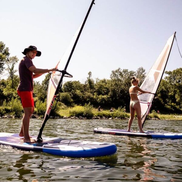 Zwei Personen surfen auf Paddelbrettern auf einem See, der von Bäumen und Grünflächen umgeben ist, unter einem klaren Himmel.