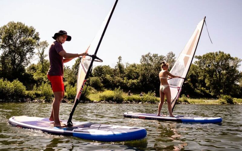 Zwei Personen surfen auf Paddelbrettern auf einem See, der von Bäumen und Grünflächen umgeben ist, unter einem klaren Himmel.