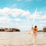 Menschen in Schwimmwesten fahren auf einem Fluss unter einem teilweise bewölkten Himmel. Im Vordergrund ist ein kleines Segelboot mit der Aufschrift "Leo" zu sehen, und links davon ein Motorboot.