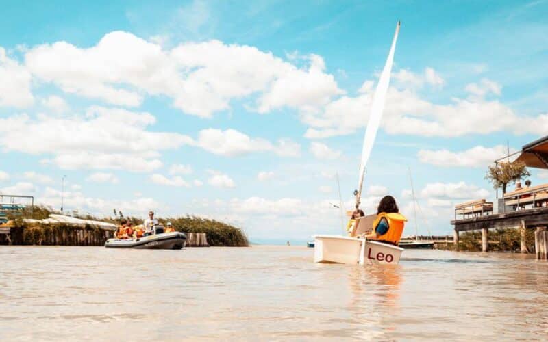 Menschen in Schwimmwesten fahren auf einem Fluss unter einem teilweise bewölkten Himmel. Im Vordergrund ist ein kleines Segelboot mit der Aufschrift "Leo" zu sehen, und links davon ein Motorboot.