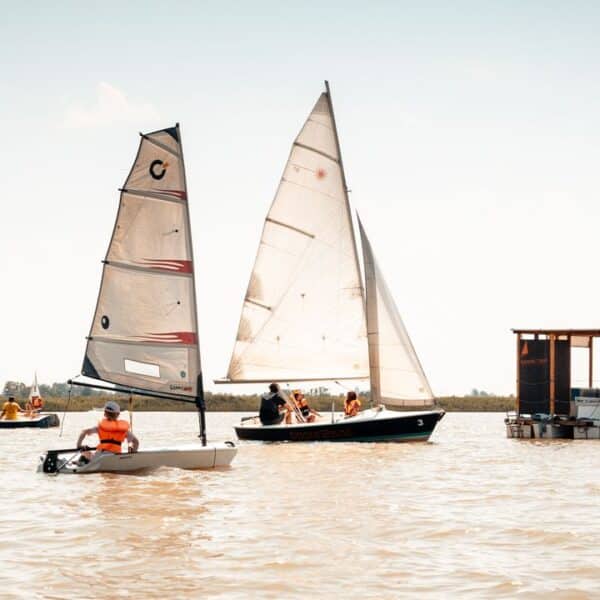 Mehrere kleine Segelboote mit Personen in Schwimmwesten und eine schwimmende Holzplattform befinden sich auf einem ruhigen Gewässer unter einem klaren Himmel.