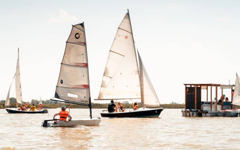 Mehrere kleine Segelboote mit Personen in Schwimmwesten und eine schwimmende Holzplattform befinden sich auf einem ruhigen Gewässer unter einem klaren Himmel.