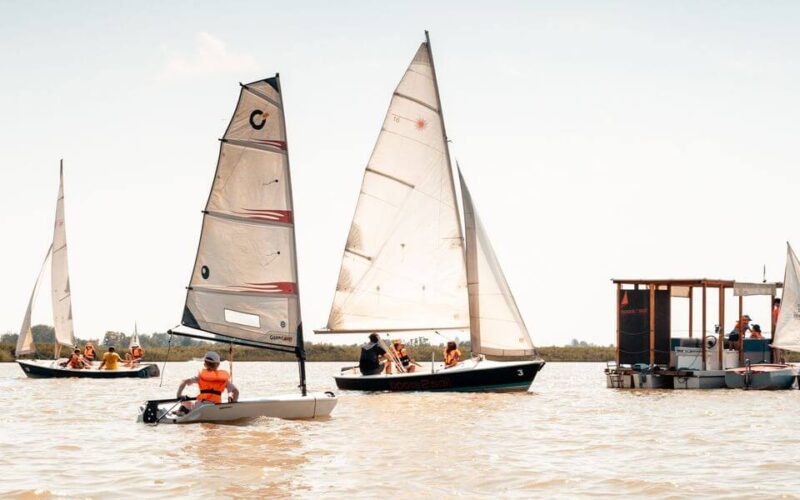 Mehrere kleine Segelboote und eine schwimmende Plattform mit Menschen befinden sich auf einem ruhigen, sonnigen Gewässer, im Hintergrund ist Land zu sehen.