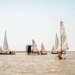 Mehrere Segelboote mit Personen in Schwimmwesten befinden sich auf einem ruhigen Gewässer unter klarem Himmel.