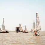 Mehrere kleine Segelboote und eine schwimmende Plattform mit Menschen auf einem ruhigen Gewässer unter einem klaren Himmel.