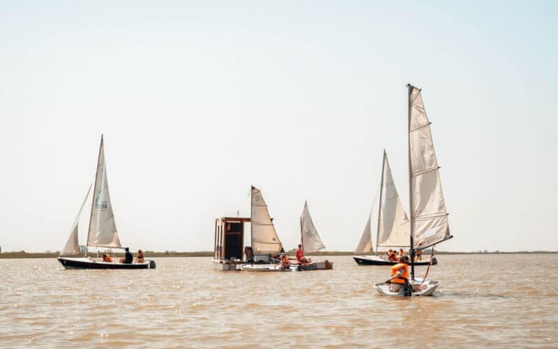 Mehrere kleine Segelboote und eine schwimmende Plattform mit Menschen auf einem ruhigen Gewässer unter einem klaren Himmel.