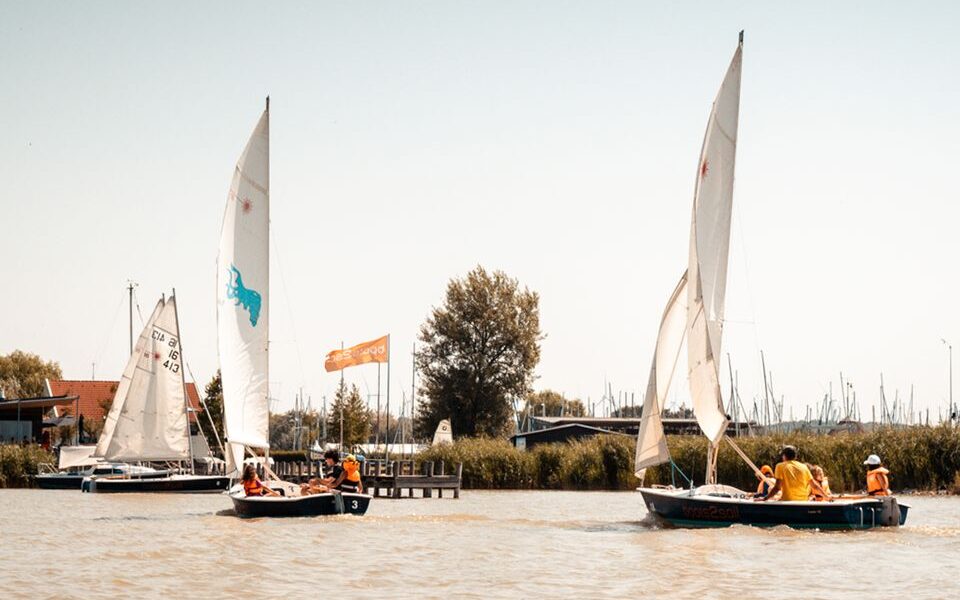 Mehrere kleine Segelboote mit Personen an Bord fahren auf einem ruhigen Gewässer in der Nähe eines Jachthafens mit einem Dock, einer Flagge und Segelbooten im Hintergrund.