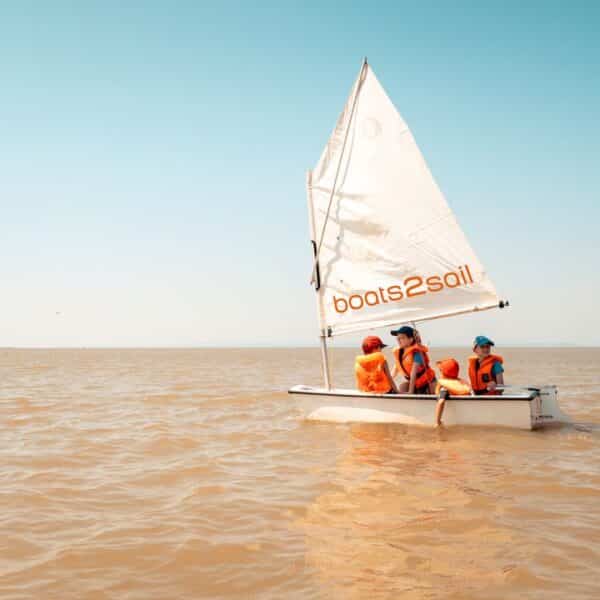 Drei Personen mit orangefarbenen Schwimmwesten sitzen in einem kleinen Segelboot mit der Aufschrift "boats2sail" auf dem ruhigen, bräunlichen Wasser unter einem klaren blauen Himmel.