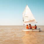 Drei Personen mit orangefarbenen Schwimmwesten segeln auf einem kleinen weißen Schlauchboot mit der Aufschrift "boats2sail" auf ruhigem, braunem Wasser unter einem klaren blauen Himmel.