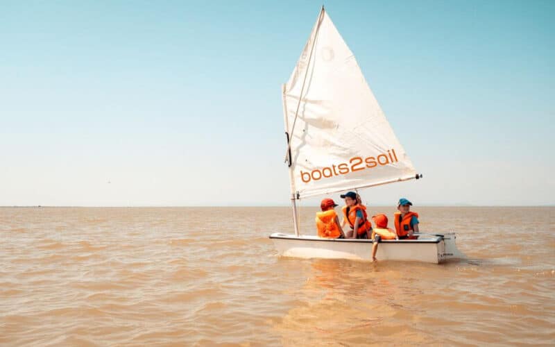 Drei Personen mit orangefarbenen Schwimmwesten segeln auf einem kleinen weißen Schlauchboot mit der Aufschrift "boats2sail" auf ruhigem, braunem Wasser unter einem klaren blauen Himmel.