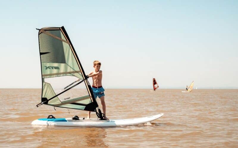 Ein Junge in Badehose surft auf ruhigem, braunem Wasser unter klarem Himmel, während in der Ferne zwei andere Windsurfer zu sehen sind.