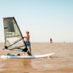 Ein Junge in Badehose steht auf einem Windsurfbrett und hält das Segel auf einem ruhigen, bräunlichen See, mit zwei anderen Windsurfern im fernen Hintergrund unter einem klaren Himmel.