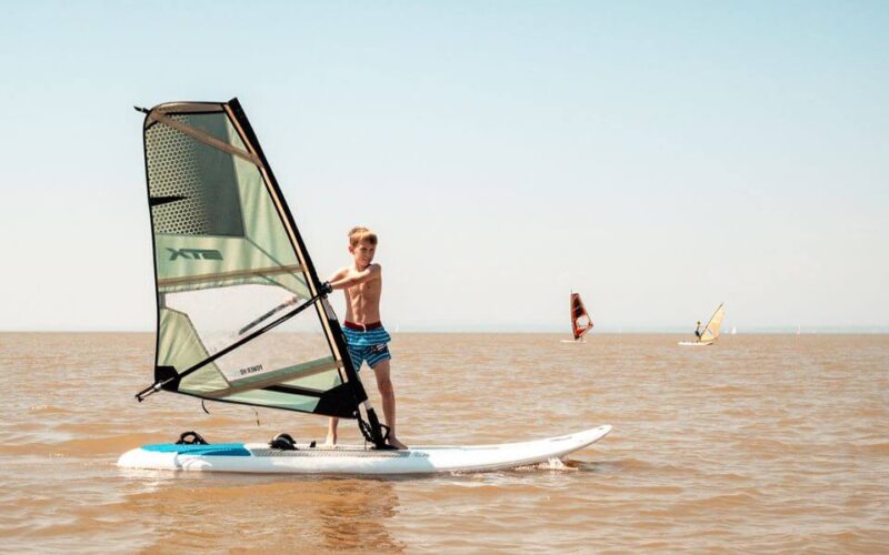 Ein Junge in Badehose steht auf einem Windsurfbrett und hält das Segel auf einem ruhigen, bräunlichen See, mit zwei anderen Windsurfern im fernen Hintergrund unter einem klaren Himmel.