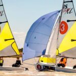 Zwei kleine Segelboote mit gelben Segeln, jeweils mit zwei Mannschaften, fahren auf einem See um die Wette. Die Segel sind mit den Nummern 2 und 3 gekennzeichnet. Der Himmel ist klar und das Wasser ist ruhig.
