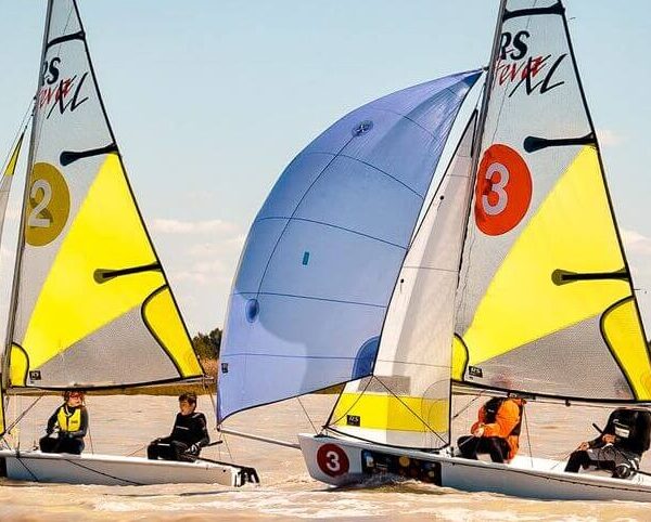 Zwei kleine Segelboote mit gelben Segeln, jeweils mit zwei Mannschaften, fahren auf einem See um die Wette. Die Segel sind mit den Nummern 2 und 3 gekennzeichnet. Der Himmel ist klar und das Wasser ist ruhig.
