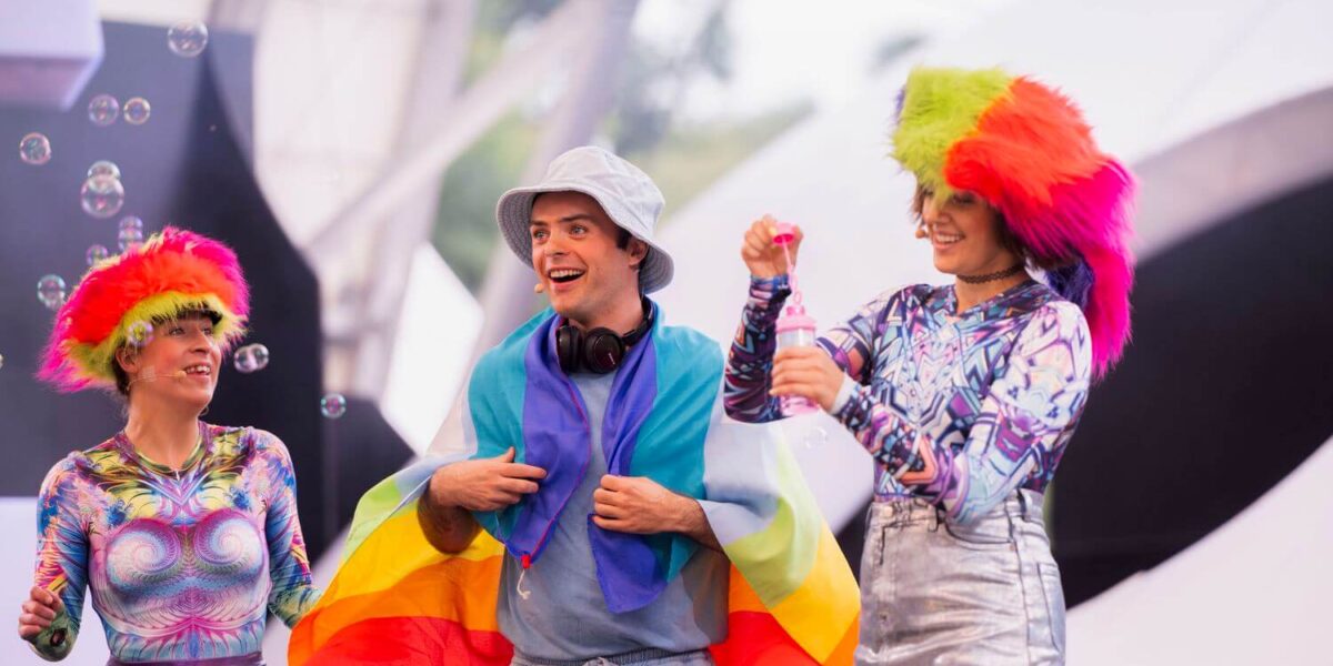 Drei Personen in farbenfrohen Outfits und mit Perücken stehen zusammen, eine Person ist in eine Regenbogenflagge gehüllt. Um sie herum schweben Seifenblasen vor einem hellen, verschwommenen Hintergrund.