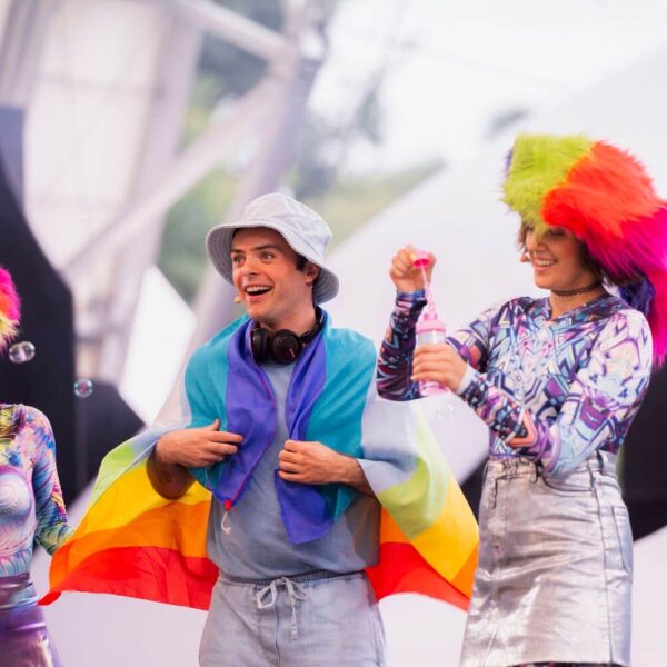 Drei Personen in farbenfrohen Outfits und mit Perücken stehen zusammen, eine Person ist in eine Regenbogenflagge gehüllt. Um sie herum schweben Seifenblasen vor einem hellen, verschwommenen Hintergrund.