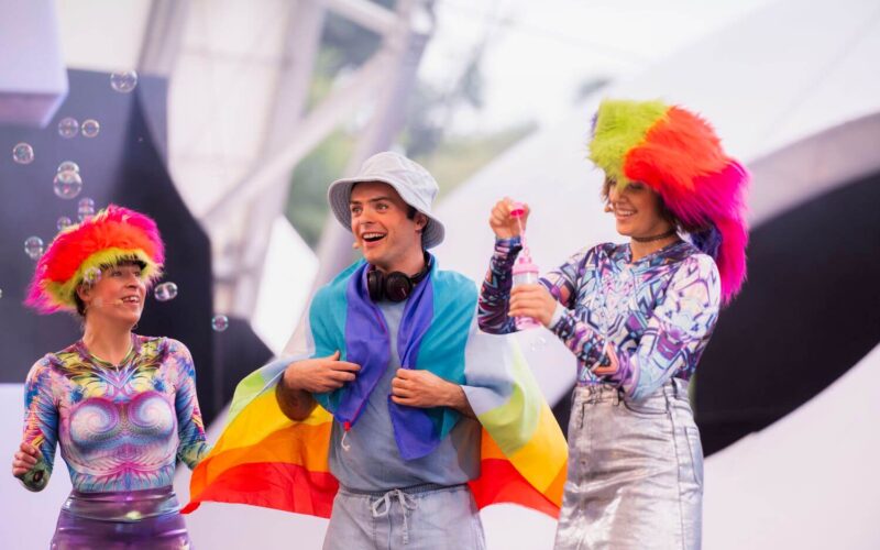 Drei Personen in farbenfrohen Outfits und mit Perücken stehen zusammen, eine Person ist in eine Regenbogenflagge gehüllt. Um sie herum schweben Seifenblasen vor einem hellen, verschwommenen Hintergrund.
