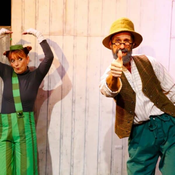 Zwei Darsteller in farbenfrohen Kostümen posieren auf der Bühne; der eine steht mit erhobenen Armen, der andere zeigt lächelnd nach vorne, vor einem Holzhintergrund.