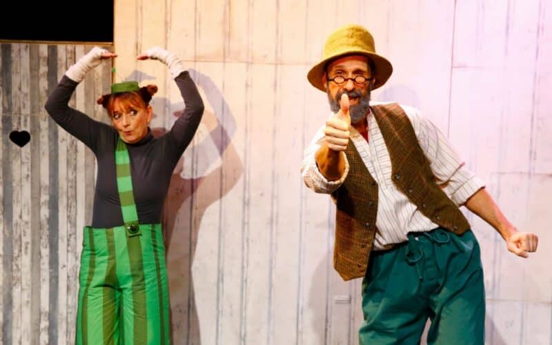 Zwei Darsteller in farbenfrohen Kostümen posieren auf der Bühne; der eine steht mit erhobenen Armen, der andere zeigt lächelnd nach vorne, vor einem Holzhintergrund.