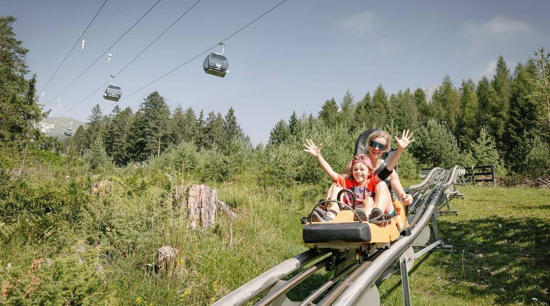 Zwei Personen fahren an einem sonnigen Tag mit einem Alpine Coaster durch ein grasbewachsenes, bewaldetes Gebiet, in dem im Hintergrund Skigondeln zu sehen sind.