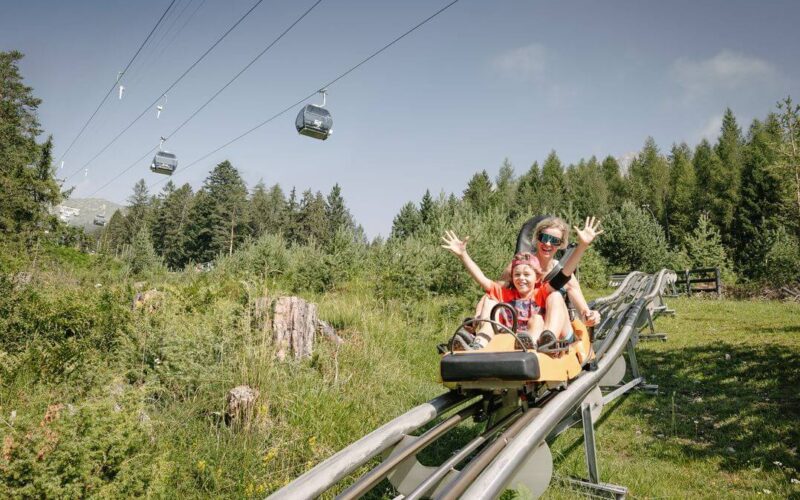 Zwei Personen fahren an einem sonnigen Tag mit einem Alpine Coaster durch ein grasbewachsenes, bewaldetes Gebiet, in dem im Hintergrund Skigondeln zu sehen sind.
