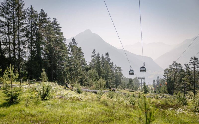 Die Sessellifte fahren über eine von Kiefern umgebene Wiese mit Bergen im Hintergrund unter einem dunstigen Himmel.