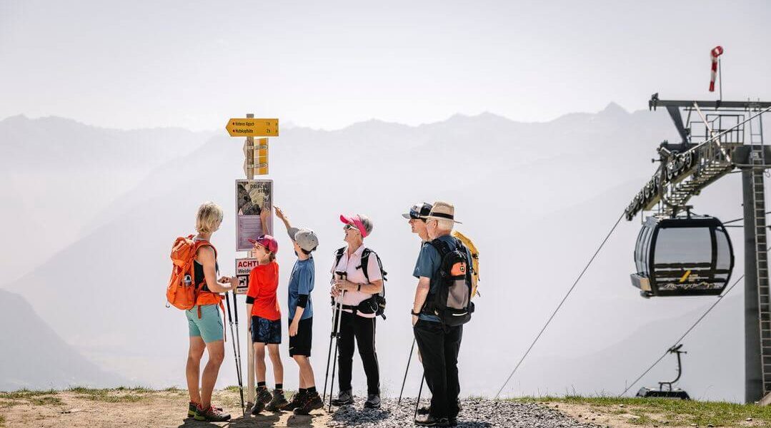 Eine Gruppe von sechs Wanderern, darunter Kinder und Erwachsene, steht in der Nähe eines Wegweisers und einer Seilbahnstation, während im Hintergrund die Berge zu sehen sind.
