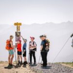 Eine Gruppe von sechs Wanderern, darunter Kinder und Erwachsene, steht in der Nähe eines Wegweisers und einer Seilbahnstation, während im Hintergrund die Berge zu sehen sind.