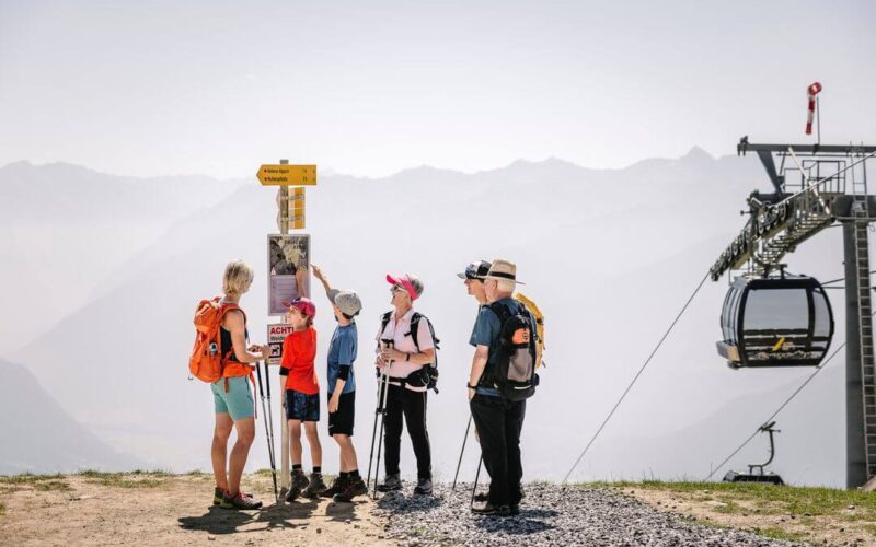 Eine Gruppe von sechs Wanderern, darunter Kinder und Erwachsene, steht in der Nähe eines Wegweisers und einer Seilbahnstation, während im Hintergrund die Berge zu sehen sind.