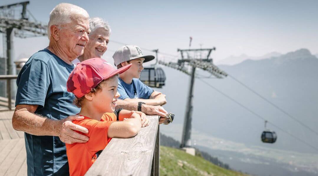 Zwei ältere Erwachsene und zwei Kinder stehen auf einem Holzdeck und blicken auf eine Berglandschaft mit Seilbahnen und Skilifttürmen im Hintergrund.