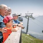 Zwei ältere Erwachsene und zwei Kinder stehen auf einem Holzdeck und blicken auf eine Berglandschaft mit Seilbahnen und Skilifttürmen im Hintergrund.