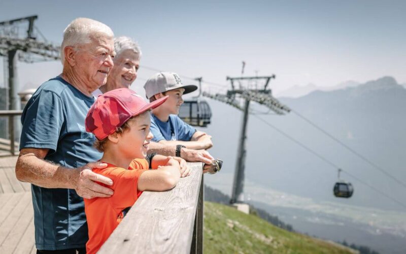 Zwei ältere Erwachsene und zwei Kinder stehen auf einem Holzdeck und blicken auf eine Berglandschaft mit Seilbahnen und Skilifttürmen im Hintergrund.