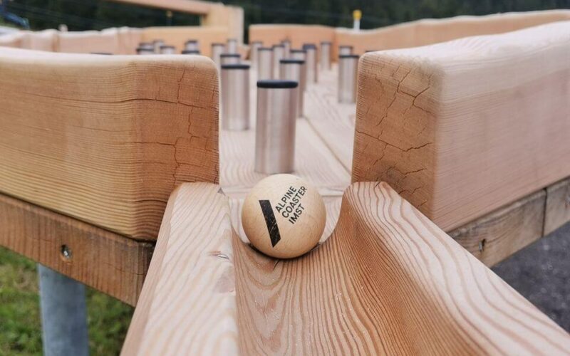 Eine Holzkugel mit der Aufschrift "Alpine Coaster MCI" steht am Anfang einer Holzbahn mit mehreren aufrecht stehenden Metallzylindern im Hintergrund.