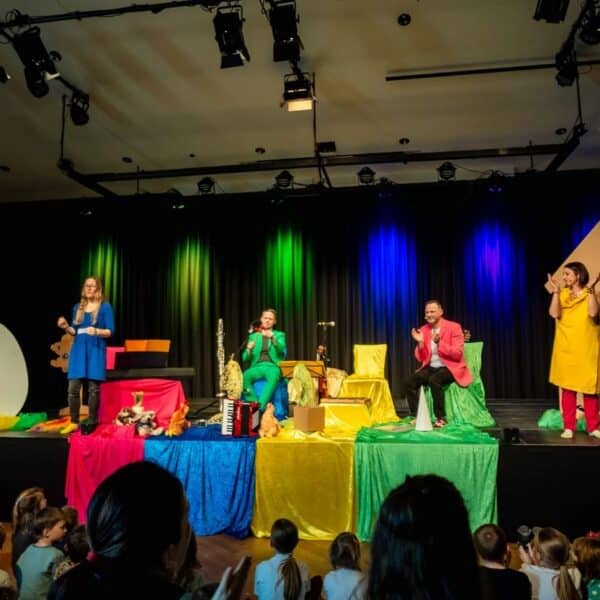 Vier Darsteller in farbenfrohen Kostümen unterhalten ein Kinderpublikum auf einer Bühne, die mit Requisiten wie einem großen Ei und einem Hausausschnitt geschmückt ist, unter Bühnenlicht.