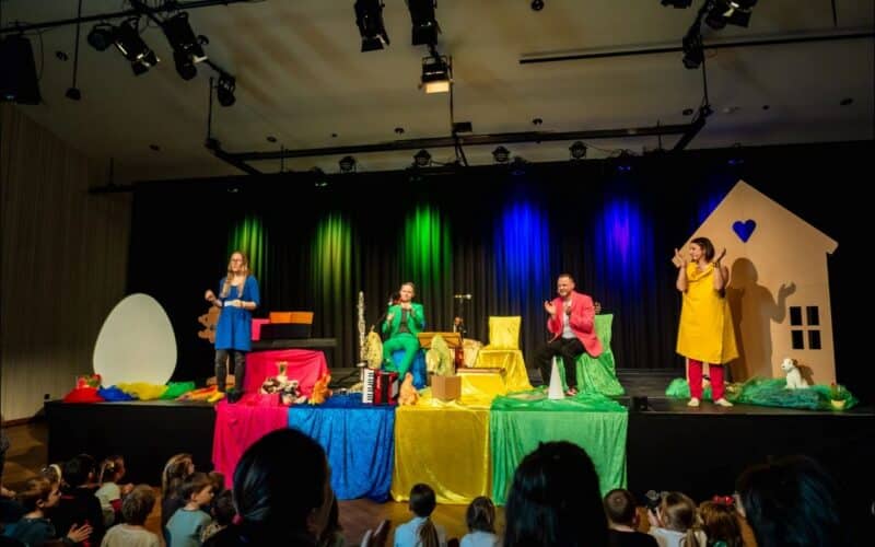Vier Darsteller in farbenfrohen Kostümen unterhalten ein Kinderpublikum auf einer Bühne, die mit Requisiten wie einem großen Ei und einem Hausausschnitt geschmückt ist, unter Bühnenlicht.
