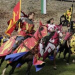 Drei Personen in mittelalterlichen Ritterkostümen reiten auf Pferden, die mit passenden heraldischen Bannern geschmückt sind, während eines Ritterturniers in der Nähe einer Steinmauer.