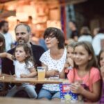Eine Familie mit Kindern sitzt an einem Tisch im Freien, isst Popcorn und sieht sich eine Veranstaltung an. Im Hintergrund sind andere Personen zu sehen.