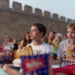 Kinder sitzen auf Decken im Freien in der Nähe einer Steinmauer, essen Snacks und trinken Getränke, während sie eine Veranstaltung oder Aufführung verfolgen.